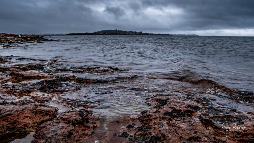Waves breaking over dark red coastal rocks under a cold, grey sky, with Churchill Island visible across the water in the distance.