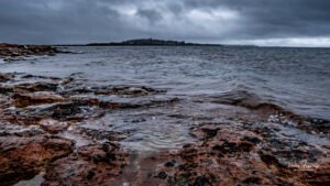 Waves breaking over dark red coastal rocks under a cold, grey sky, with Churchill Island visible across the water in the distance.